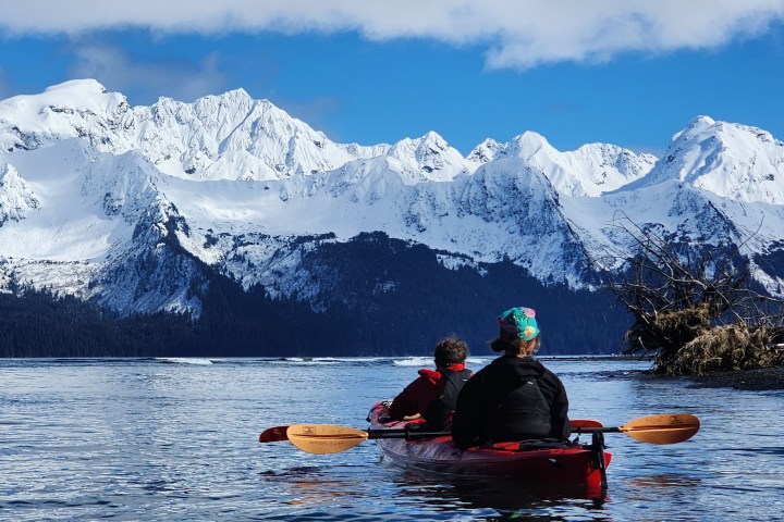 Two people kayaking towards snowy mountains on a clear day.