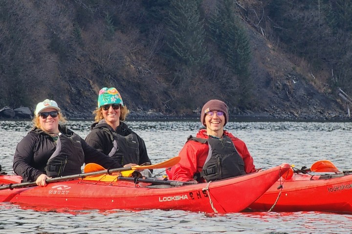 Three people kayaking on a lake in red kayaks, wearing jackets and hats.