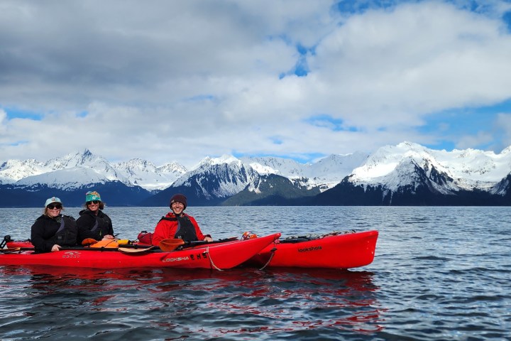 Three people kayaking on a lake with snow-capped mountains in the background.