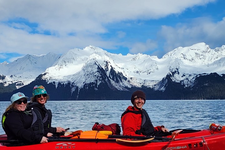 Three people kayaking on a lake with snow-capped mountains in the background.