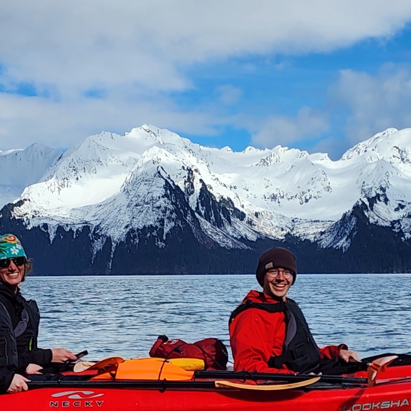 Three people kayaking on a lake with snow-capped mountains in the background.