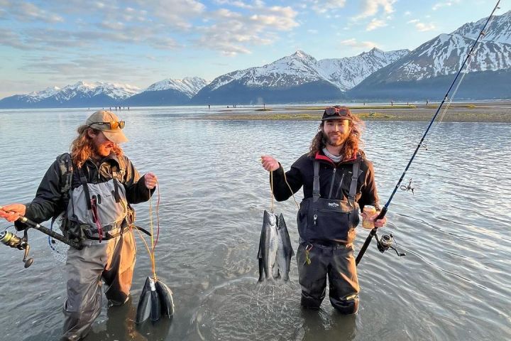 Two people stand in water holding fish and fishing rods, with snow-capped mountains in the background.