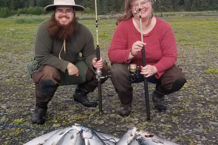Two people with fishing rods kneel next to a pile of fish with snowy mountains in the background.