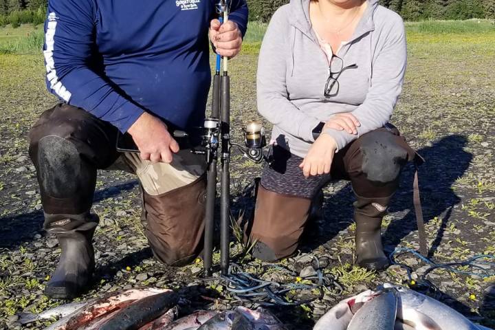Two people kneeling on gravel with freshly caught fish and snow-capped mountains in the background.