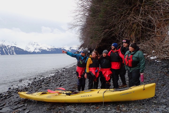 Group in lifejackets on rocky shore next to yellow kayak, snowy mountains in background.