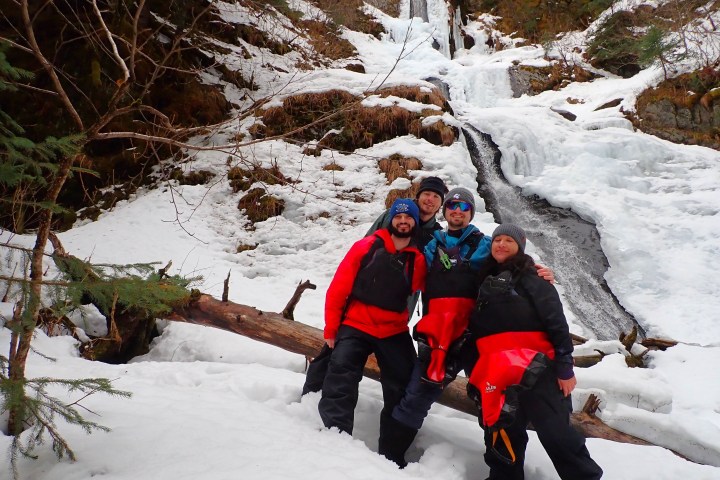 Four people in winter gear stand on a snowy hill in front of a frozen waterfall.