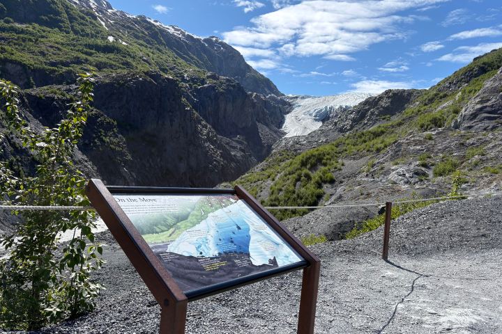 Informational sign with mountainous landscape and distant glacier under a blue sky.