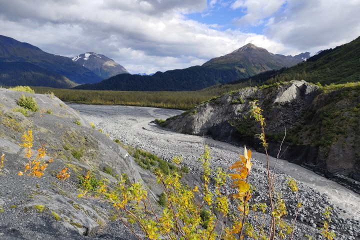 Mountains, riverbed, and autumn foliage under a partly cloudy sky.