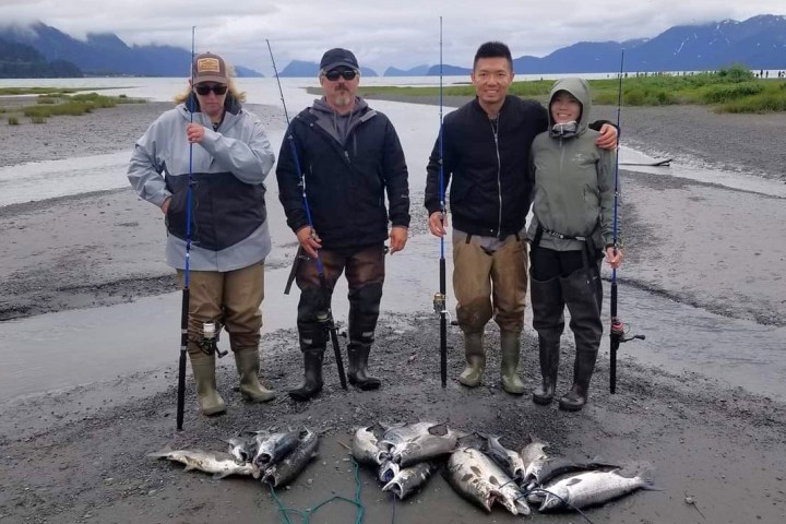 Four people with fishing rods and salmon by a river, wearing outdoor gear.