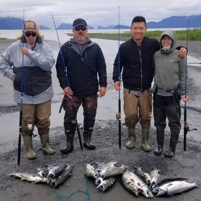 Four people with fishing rods and salmon by a river, wearing outdoor gear.