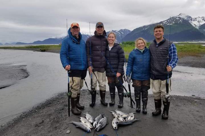 Five people in fishing gear stand on a riverbank with several fish on the ground, mountains in the background.