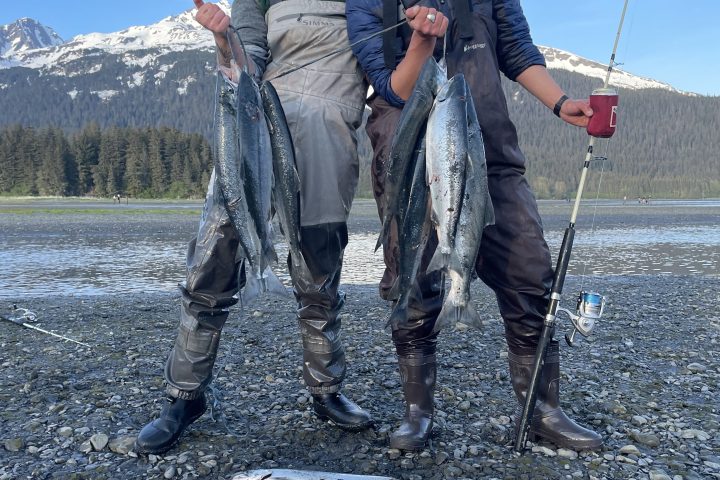 Two people in fishing gear holding fish, standing on rocky shore with mountains in the background.