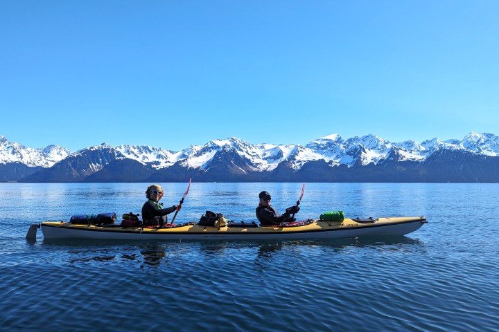 Two people kayaking on a calm lake with snow-capped mountains in the background.