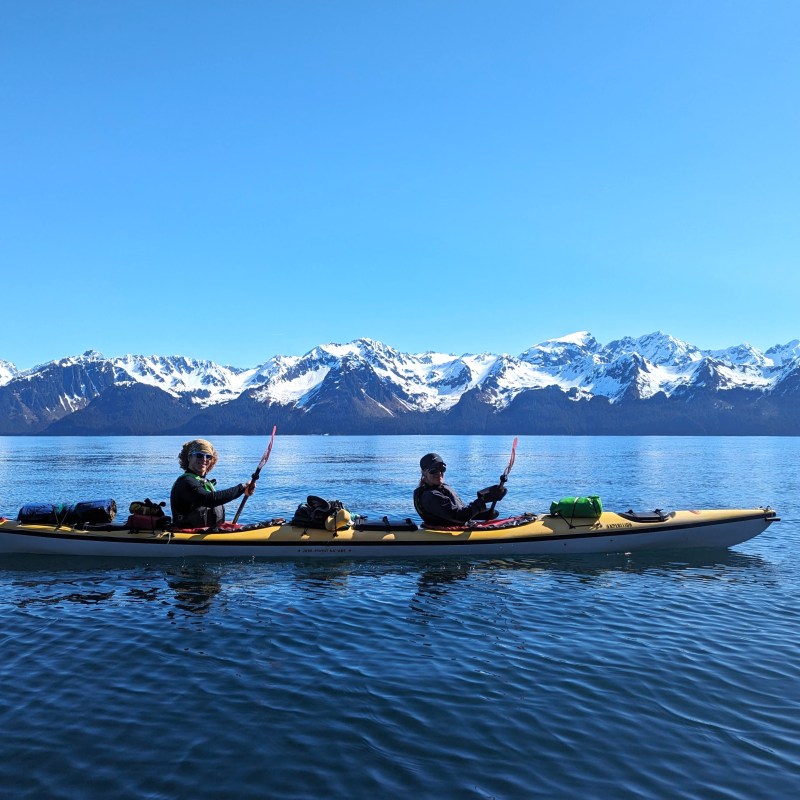 Two people kayaking on a calm lake with snow-capped mountains in the background.