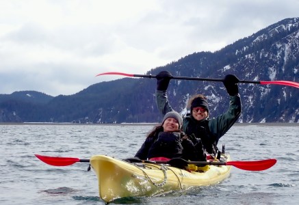 Two people kayaking on a lake with mountains in the background, one holding a paddle above their head.