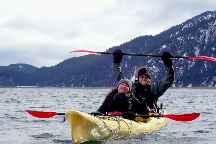 Two people kayaking on a lake with mountains in the background, one holding a paddle above their head.