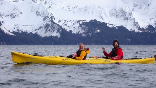Two people in a yellow kayak on a lake with snowy mountains in the background.