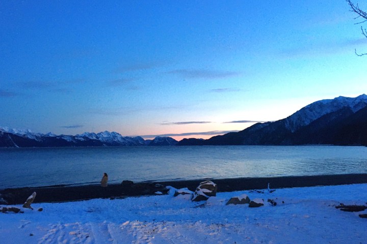 Snowy beach at dusk with mountains and a calm lake under a clear sky.