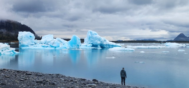 Person standing by a lake with blue icebergs and mountains under a cloudy sky.