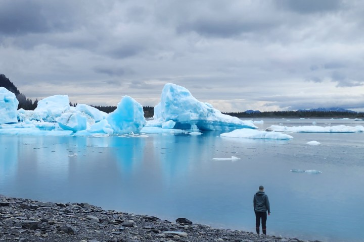 Person standing by a lake with blue icebergs and mountains under a cloudy sky.
