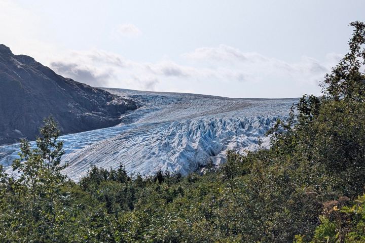 Large glacier between rocky mountains and green foliage under a clear sky.