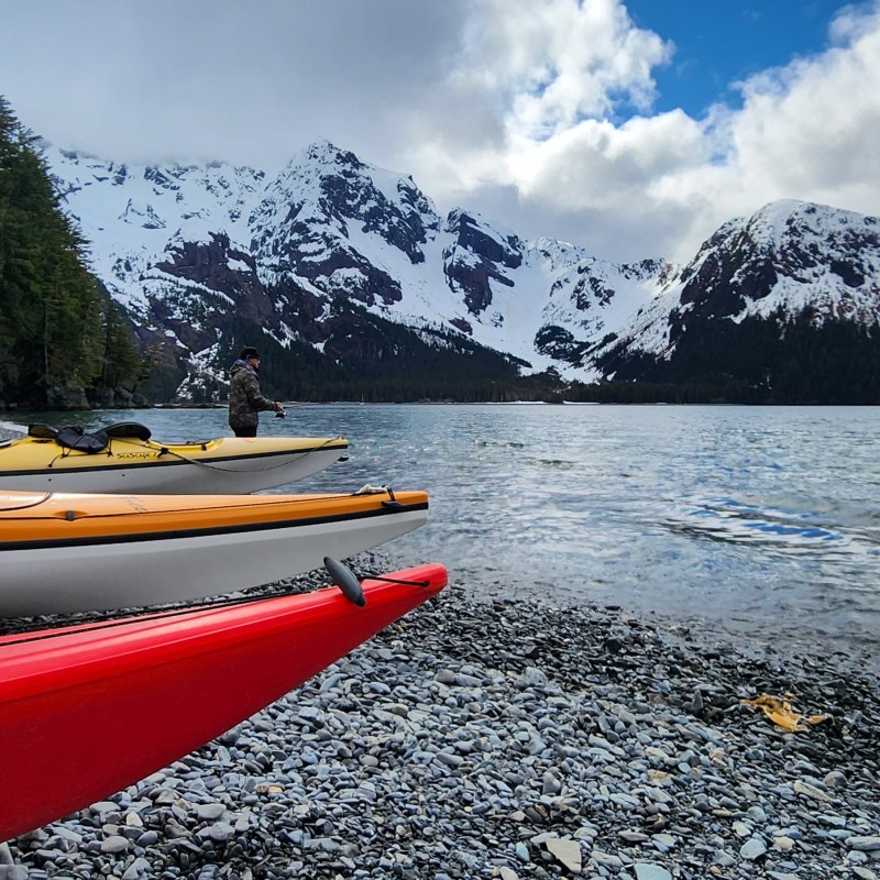 Colorful kayaks on rocky shore with snowy mountains and cloudy sky in the background.