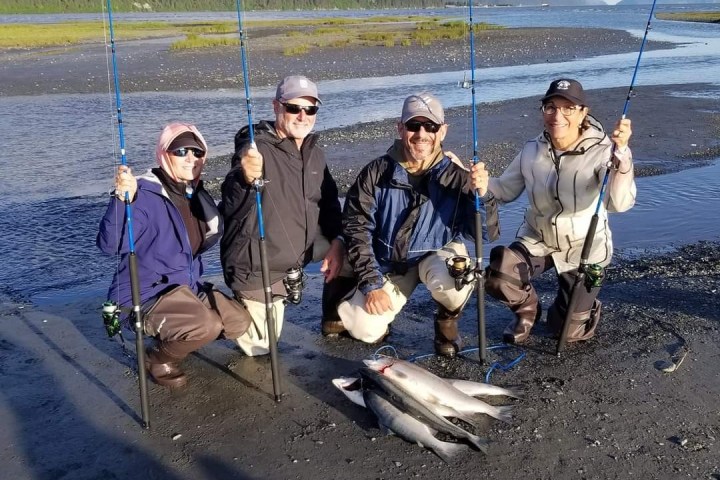 Four people in waders smiling with fishing rods and caught fish by a river.