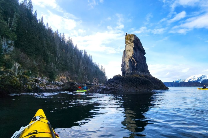 Kayaks on calm water near a tall rock formation and forested shoreline under a blue sky.