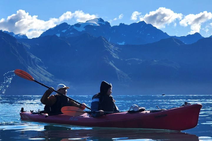 Two people kayaking in a red kayak on a lake with mountainous backdrop under a blue sky.