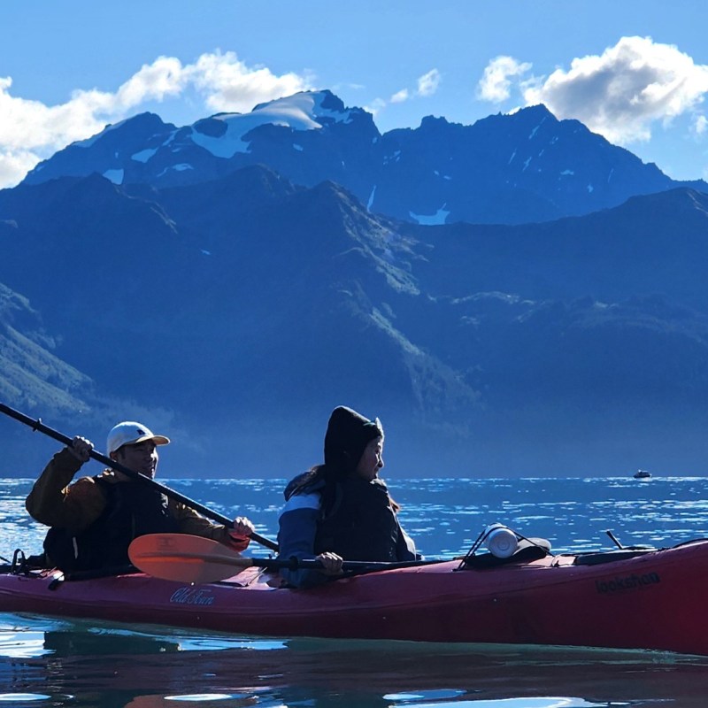 Two people kayaking in a red kayak on a lake with mountainous backdrop under a blue sky.