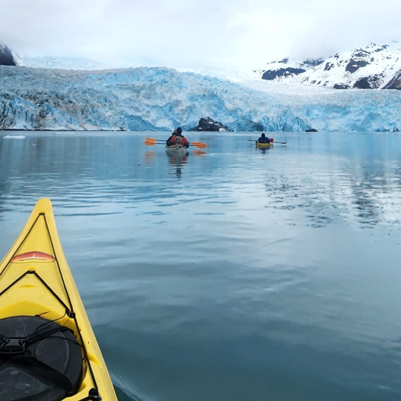 Kayakers paddle towards a large glacier surrounded by snow-capped mountains.