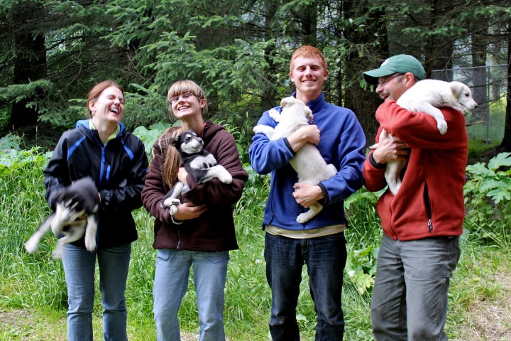 Four people holding puppies, standing in a forest, all smiling and laughing.