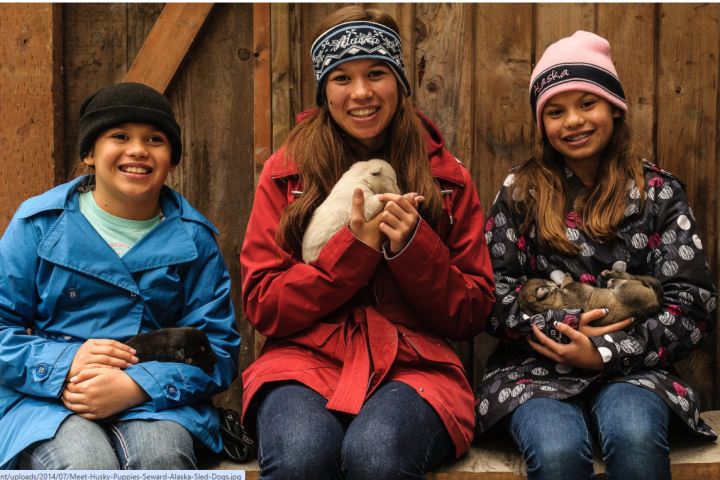 Three girls in jackets hold puppies, smiling in front of a wooden wall.