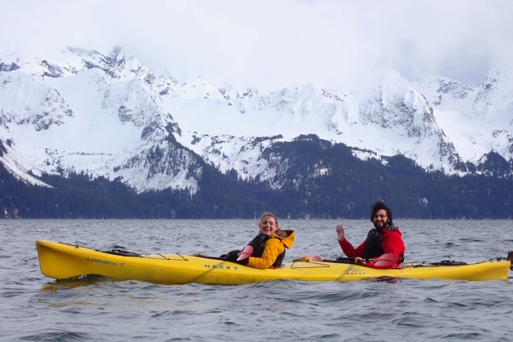 Two people kayaking on a lake with snow-covered mountains in the background.