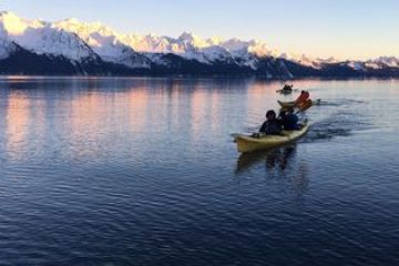 Two people kayaking on a calm lake with snow-capped mountains in the background at sunset.