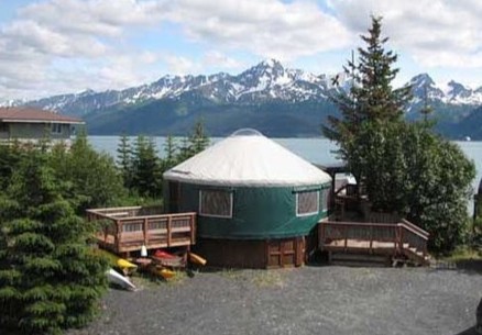 Round yurt with wooden deck, surrounded by trees, mountains in distance.