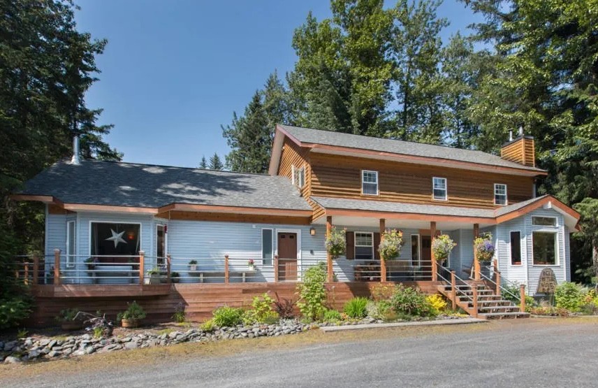 Two-story house with wood siding, porch, and flower pots, surrounded by trees.