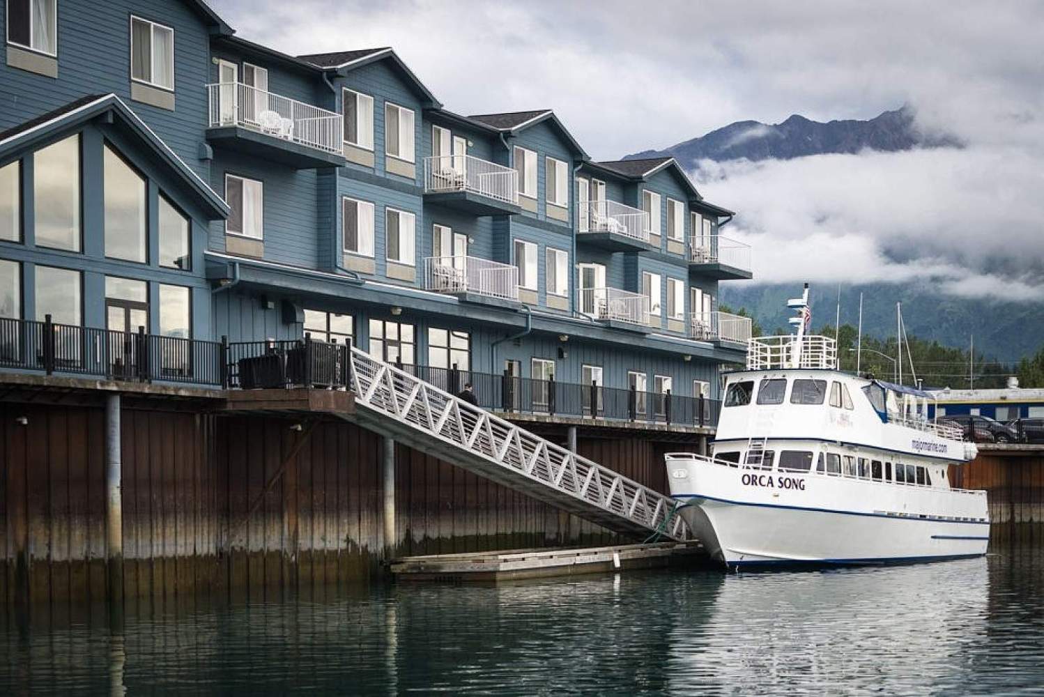 Boat named Orca Song docked beside a blue multi-story building with mountains in the background.