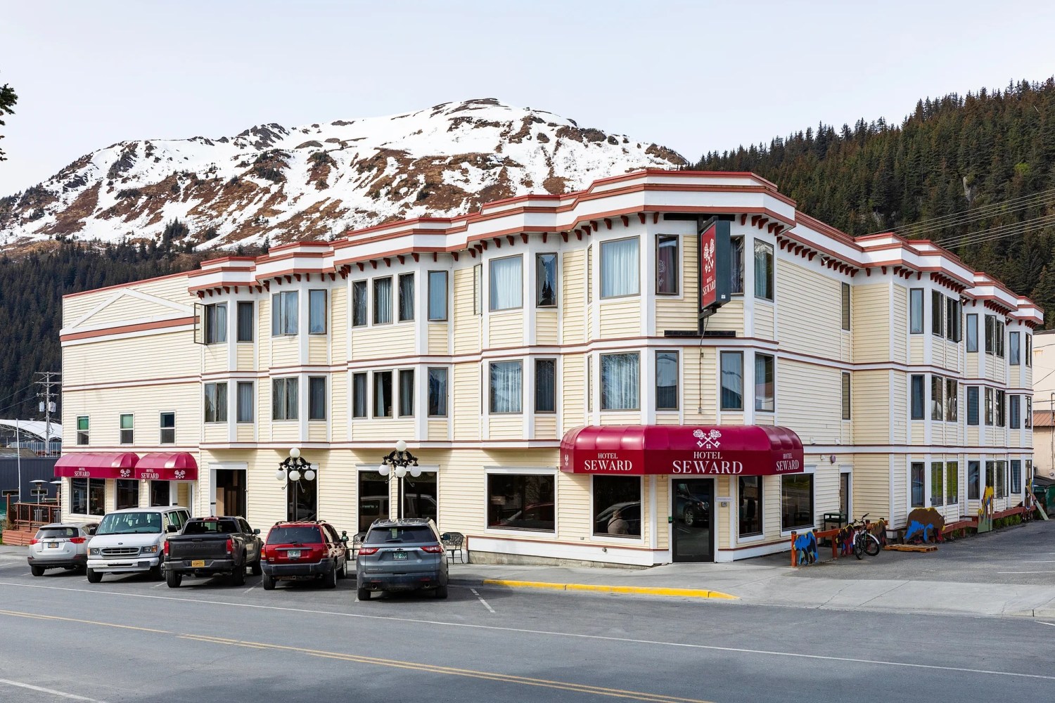Three-story hotel with red awnings and mountain backdrop, parked cars in front.