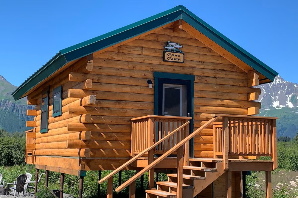 A wooden cabin on stilts with a green roof, nestled in a mountainous area with clear skies.