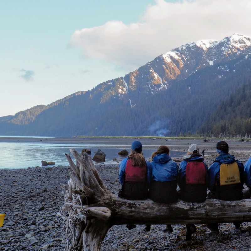 People in blue jackets sitting on a log by a rocky shore with mountains in the background.