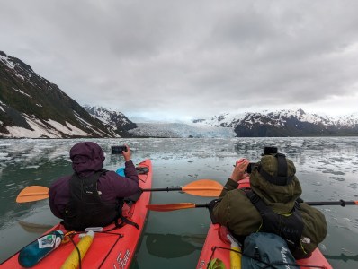 Two kayakers photographing a glacier surrounded by snowy mountains on a cloudy day.