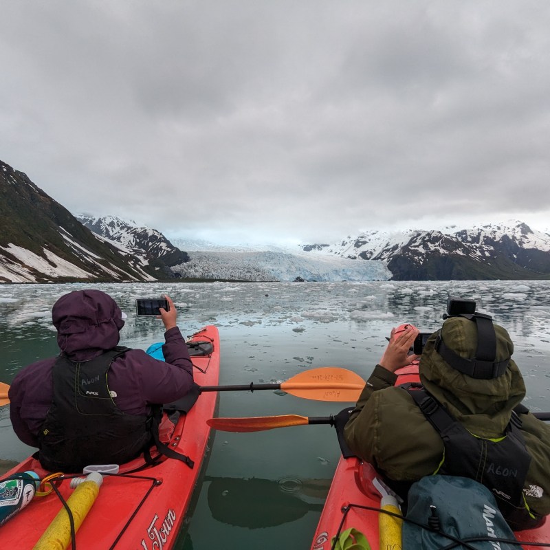 Two kayakers photographing a glacier surrounded by snowy mountains on a cloudy day.