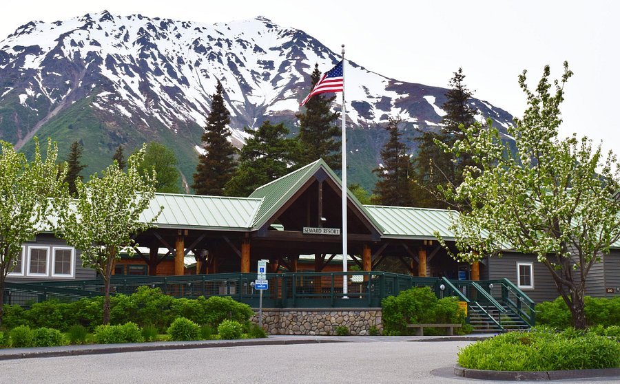 Mountain lodge with green roof, US flag, trees, and snowy peak backdrop.