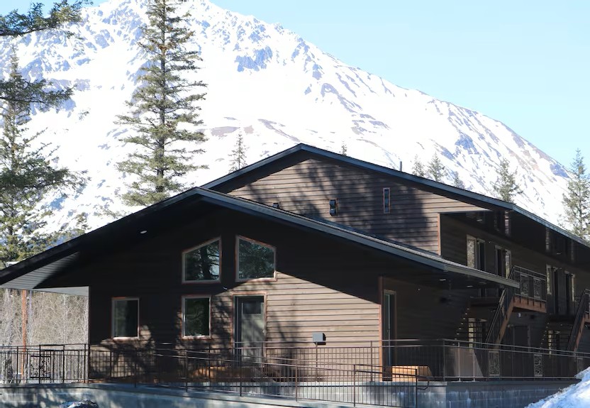 Wooden cabin with snowy mountain backdrop and pine trees.