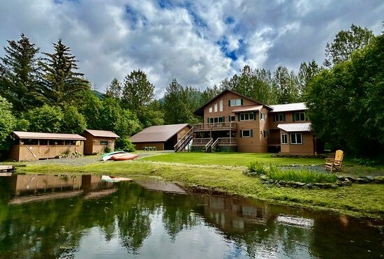 Large house with outbuildings by a pond, surrounded by trees under a cloudy sky.