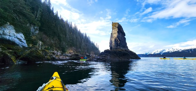 Kayakers near rocky outcrop with forest and snowy mountains in background.