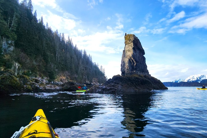 Kayakers near rocky outcrop with forest and snowy mountains in background.