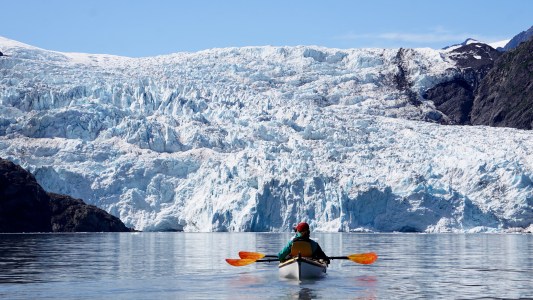 Person kayaking towards a large glacier under clear blue sky.