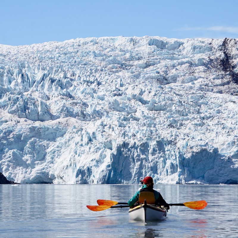 Person kayaking towards a large glacier under clear blue sky.